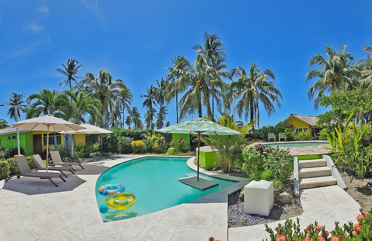 A spacious pool area is surrounded by lush palm trees and colorful gardens. Lounge chairs are situated near the water, and umbrellas provide shade. Vibrant beach floats can be seen in the clear pool, enhancing the inviting outdoor atmosphere.
