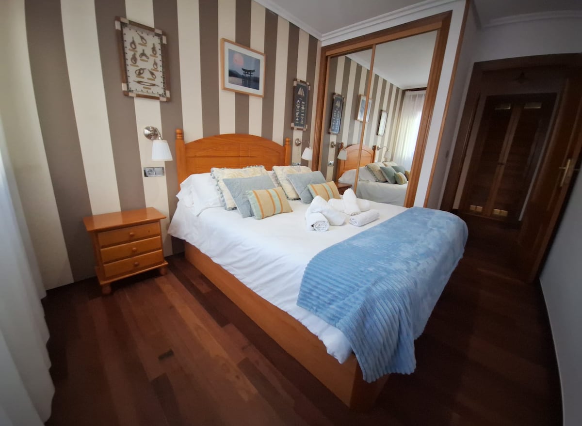 A cozy bedroom features a spacious bed adorned with soft pillows and a light blue throw. A wooden nightstand sits beside the bed, and a full-length mirror reflects the room. Subtle striped wallpaper complements the warm wooden flooring.