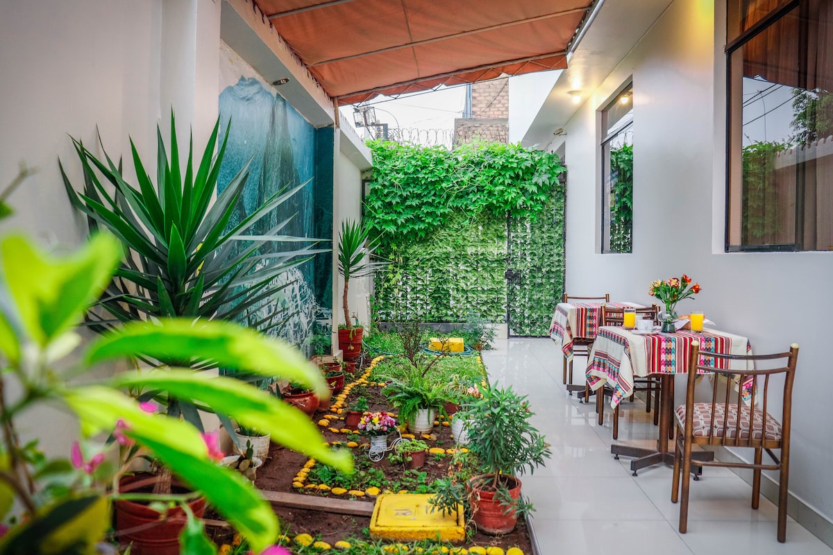A vibrant outdoor space is presented, featuring a garden area with various plants and flowers. Two tables adorned with colorful tablecloths are situated under a covered roof, providing a comfortable spot to enjoy meals. Greenery lines the walls, adding a refreshing touch to the ambiance.