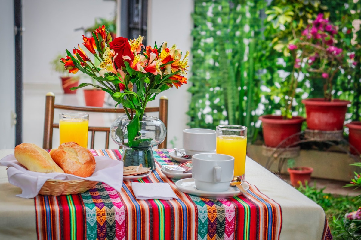 A dining table is set with a colorful woven tablecloth, featuring a vase of fresh flowers. Two glasses of orange juice and two white cups are placed alongside a basket of bread rolls. Lush greenery and potted plants create a vibrant backdrop.