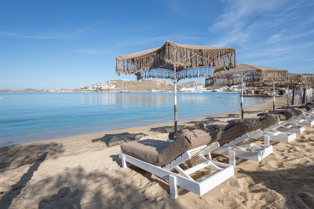 A serene beach scene features sun loungers arranged under stylish umbrellas, placed on soft sand. The calm sea extends into the horizon, reflecting blue skies. Nearby, a coastal landscape is visible, enhancing the tranquil atmosphere of this beach setting.