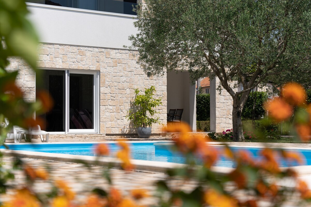 A clear view of a refreshing outdoor pool is observed through lush, blooming foliage. The pool area is framed by a stone facade of the building, with large glass doors leading inside. Comfortable seating and greenery enhance the tranquil setting.