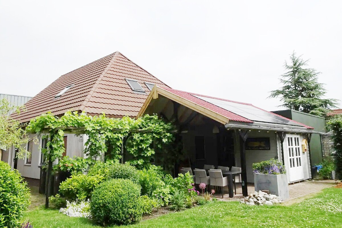 The exterior of the B&B features a sloped roof with a distinctive overhang supported by wooden beams. A patio area with a table and chairs is surrounded by lush greenery, including climbing plants and neatly trimmed shrubs, creating an inviting outdoor space.