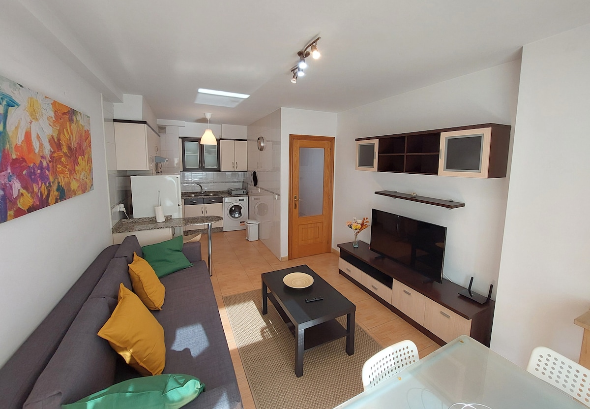 An open-plan living area features a sofa with decorative cushions, a coffee table, and a dining table. A modern kitchen is seen in the background, equipped with essential appliances. Natural light enters through a window, enhancing the inviting feel of the space.