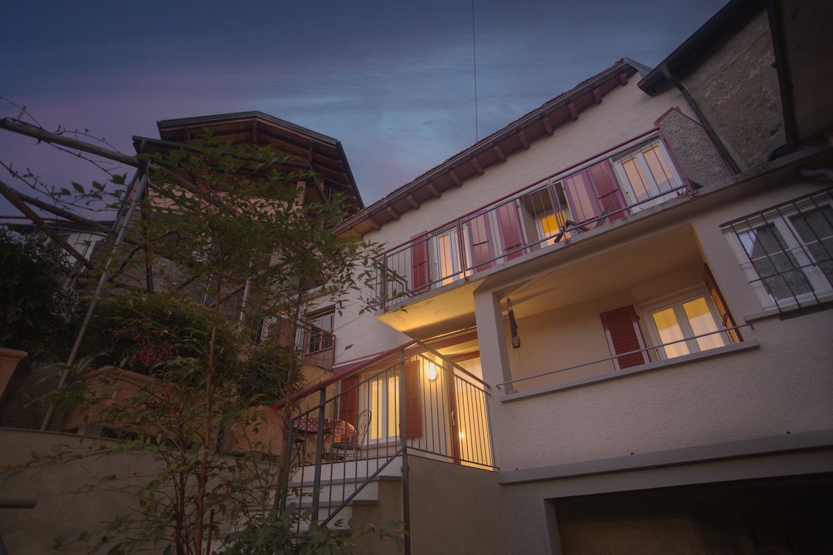 The exterior of the house is showcased in evening light, highlighting a charming blend of traditional architecture with modern updates. Soft glow from interior lights suggests warmth inside, while balcony railings and shutters add to its inviting character. Surrounding greenery provides a natural touch.