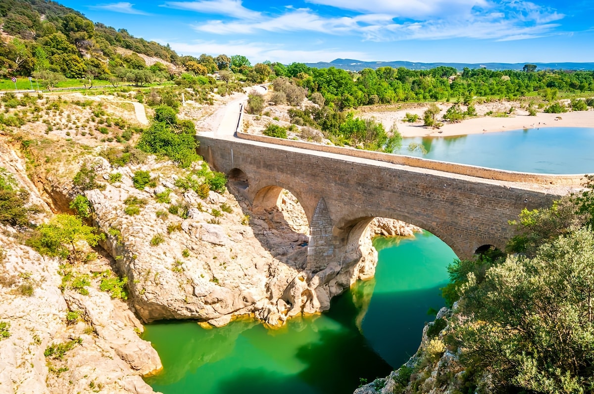 A stone bridge arches gracefully over a clear, turquoise river, flanked by rugged rock formations. Lush greenery surrounds the water’s edge, with rolling hills in the background under a bright blue sky. The scene conveys a sense of tranquility and natural beauty.