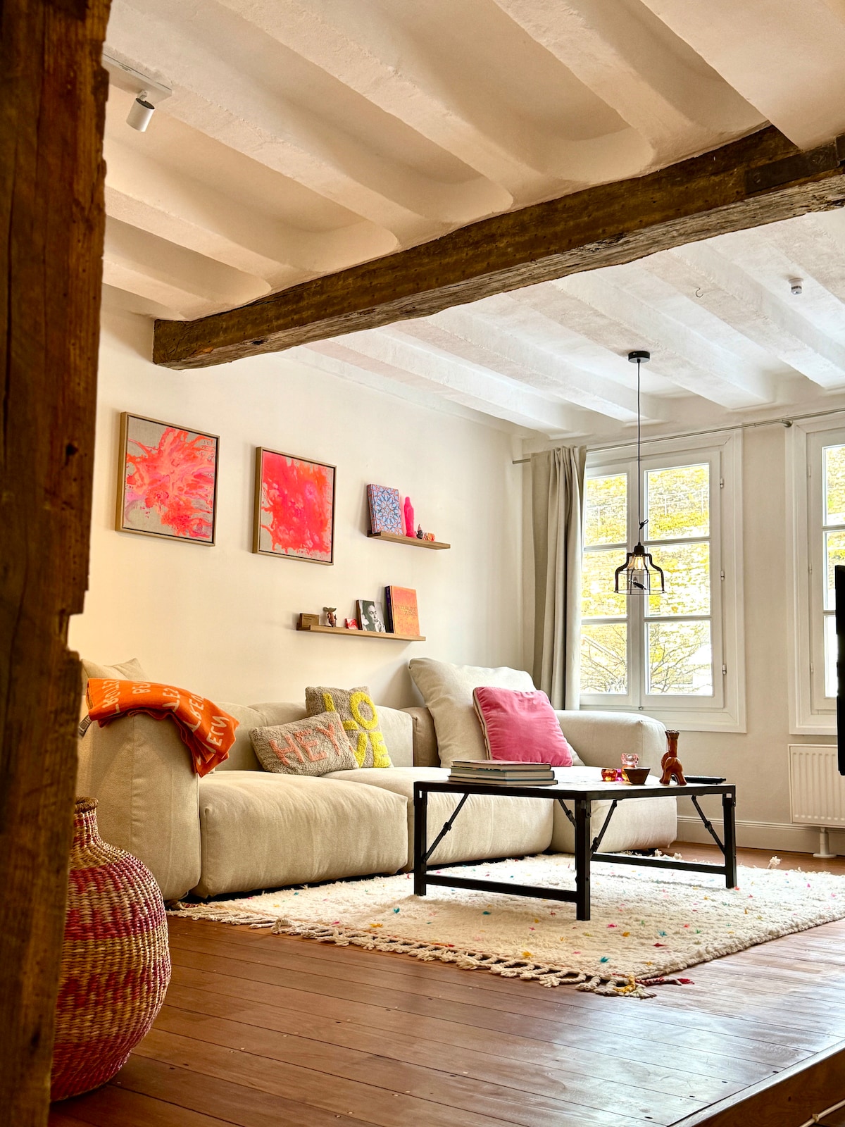The living area features a neutral-toned sofa adorned with pink and orange pillows, positioned across from a simple black coffee table. Colorful artwork is displayed on the light walls, complemented by natural light streaming through large windows. A textured rug lies beneath, adding warmth to the space.
