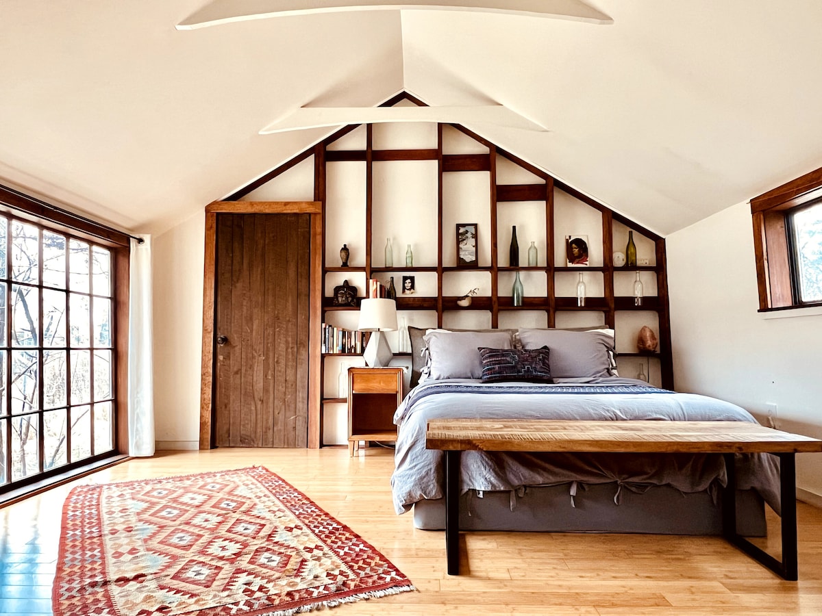 A bright sleeping area features a spacious bed against a backdrop of a sloped ceiling. A wooden bench sits at the foot of the bed, with a patterned rug laid beneath. Shelves display various decorative items, and natural light floods in from large windows.
