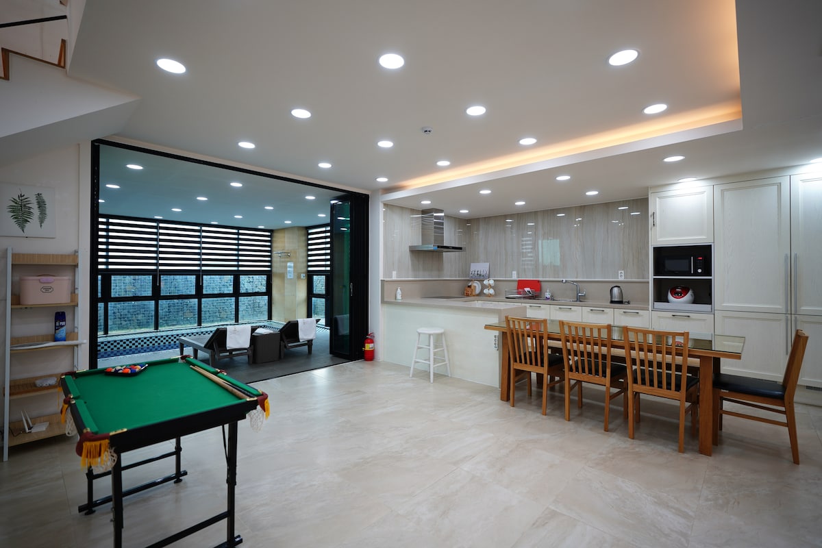 A bright and spacious kitchen and dining area features modern appliances and a large dining table with wooden chairs. Adjacent to the kitchen is a game table, ideal for indoor activities. Large glass doors allow natural light to fill the room and provide views of the outdoor space.