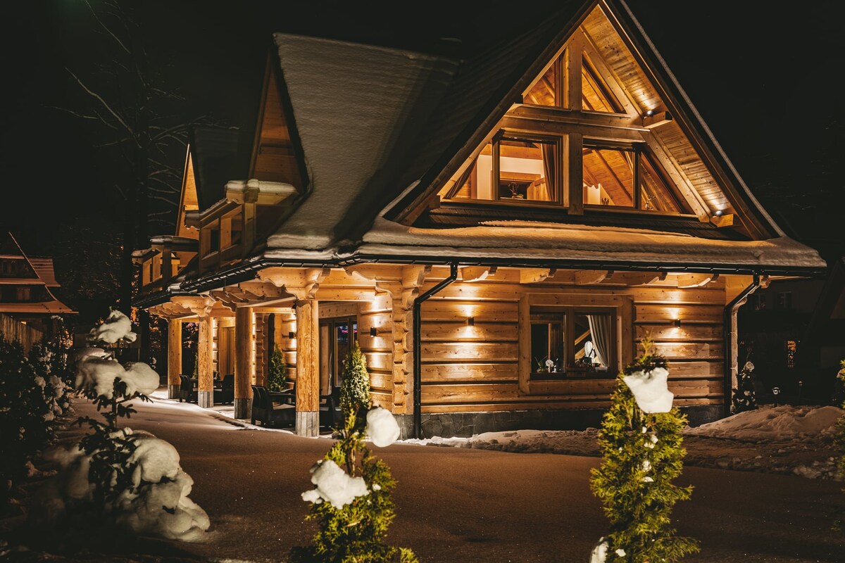 The exterior of a wooden chalet is illuminated at night, with warm lights enhancing its architectural features. Snow blankets the ground and rooftops, while decorative trees flank the path leading to the entrance. The structure showcases a combination of traditional and modern alpine design.