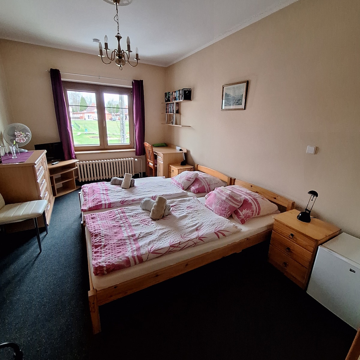 A spacious room features a double bed with soft linens and two decorative towels neatly arranged. Natural light enters through a window, complemented by curtains. A wooden dresser and a small table with a chair are present, along with a compact fridge and additional shelves.