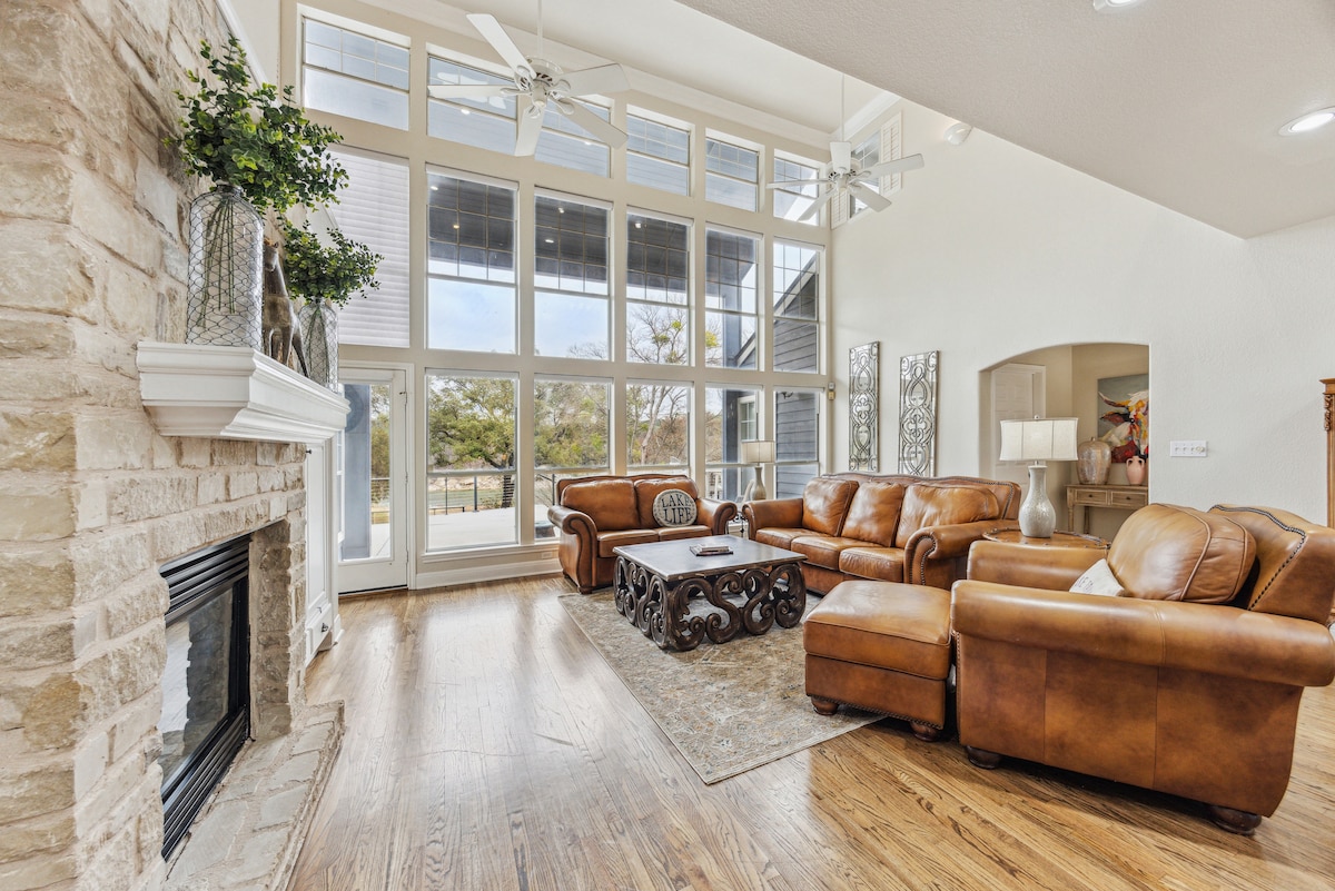 A welcoming living area features large windows, allowing natural light to fill the space. Two brown leather sofas are situated around a decorative coffee table, while a stone fireplace adds a touch of warmth. Light wood flooring enhances the overall spacious ambiance.