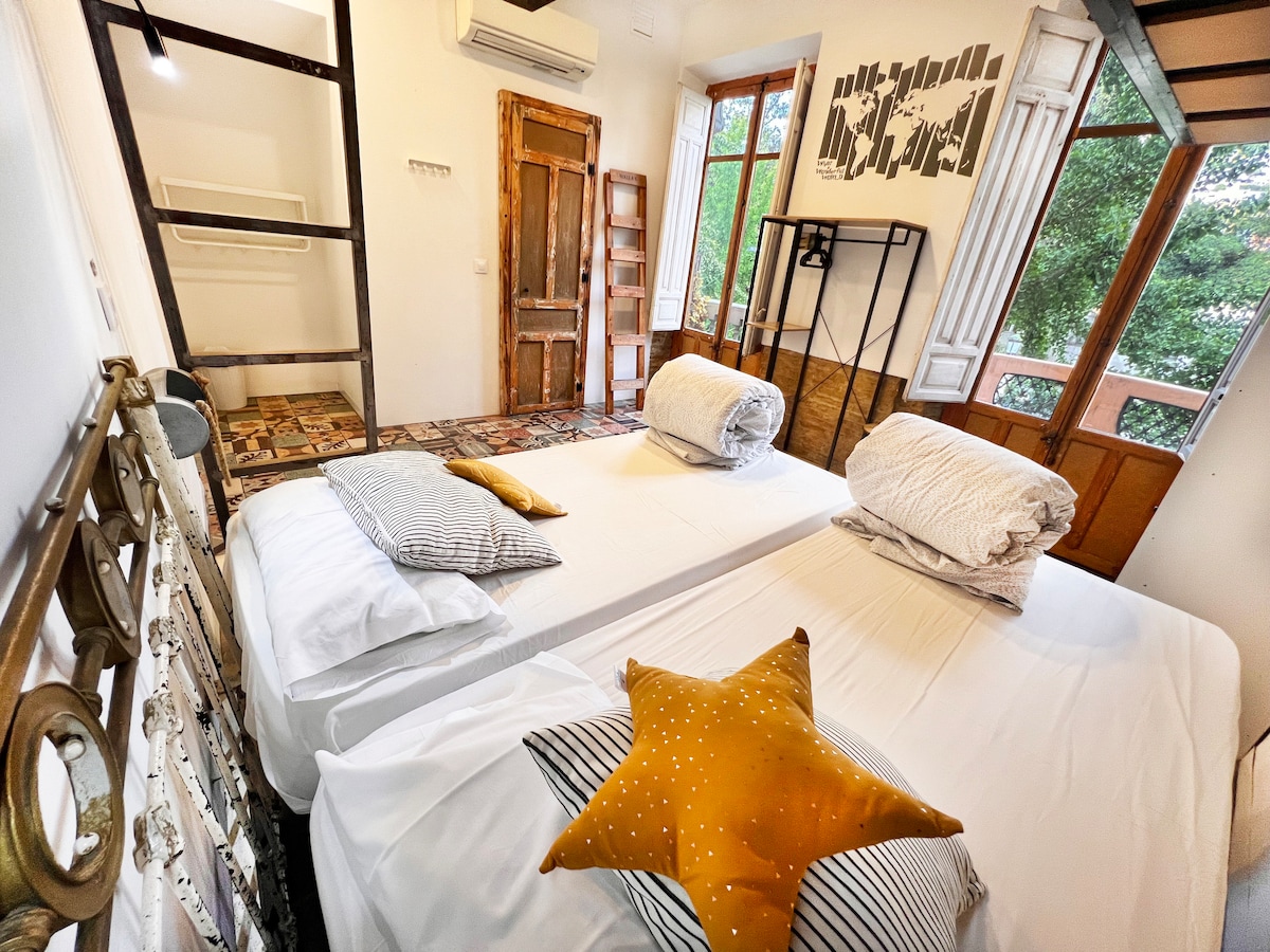 The room features two spacious beds with light linens and decorative pillows, including a star-shaped pillow. Large windows provide natural light and views, while wooden accents and a mix of textures create a comfortable and inviting space. A bookshelf and open wardrobe are also present.