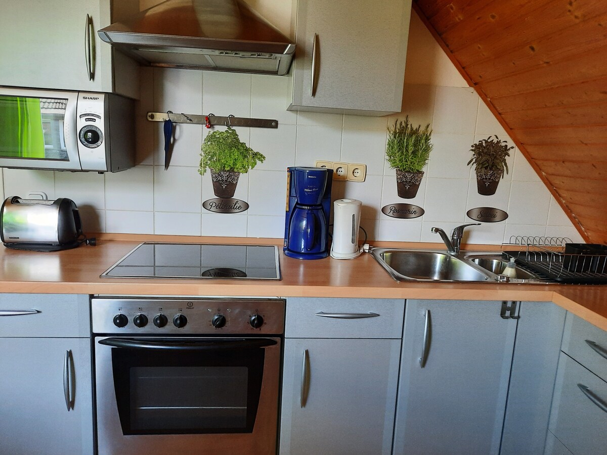 The kitchen features modern appliances, including an oven, coffee maker, and toaster. White tiles adorn the walls, accented by decorative plant motifs. A stainless steel sink and ample countertop space are available for meal preparation, complemented by a sleek design and practical cabinetry.