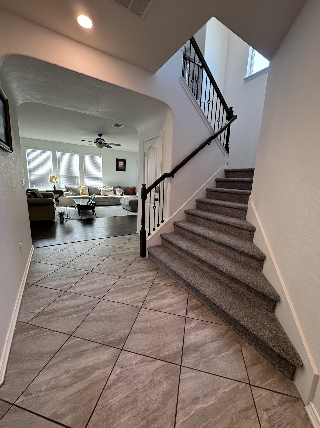 A well-lit entryway features a staircase with a black iron railing leading to an upper level. The tiled floor showcases a contemporary design, while an arched opening reveals a bright living area with large windows and comfortable seating visible in the background.