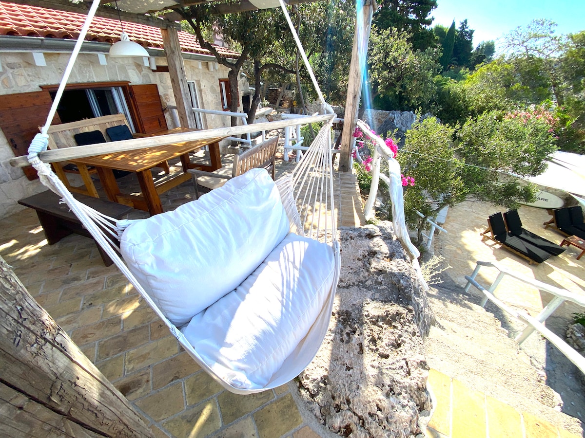A comfortable hanging hammock is positioned under a shaded pergola, featuring a plush white cushion for relaxation. A wooden dining table and chairs are visible in the background, surrounded by lush greenery and vibrant floral accents.