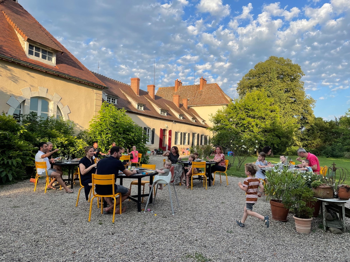 An outdoor dining area is set amid greenery, showcasing several tables with guests enjoying meals. Colorful chairs surround the tables, and children can be seen playing nearby. The backdrop features a charming stone building and a large tree under a partly cloudy sky.