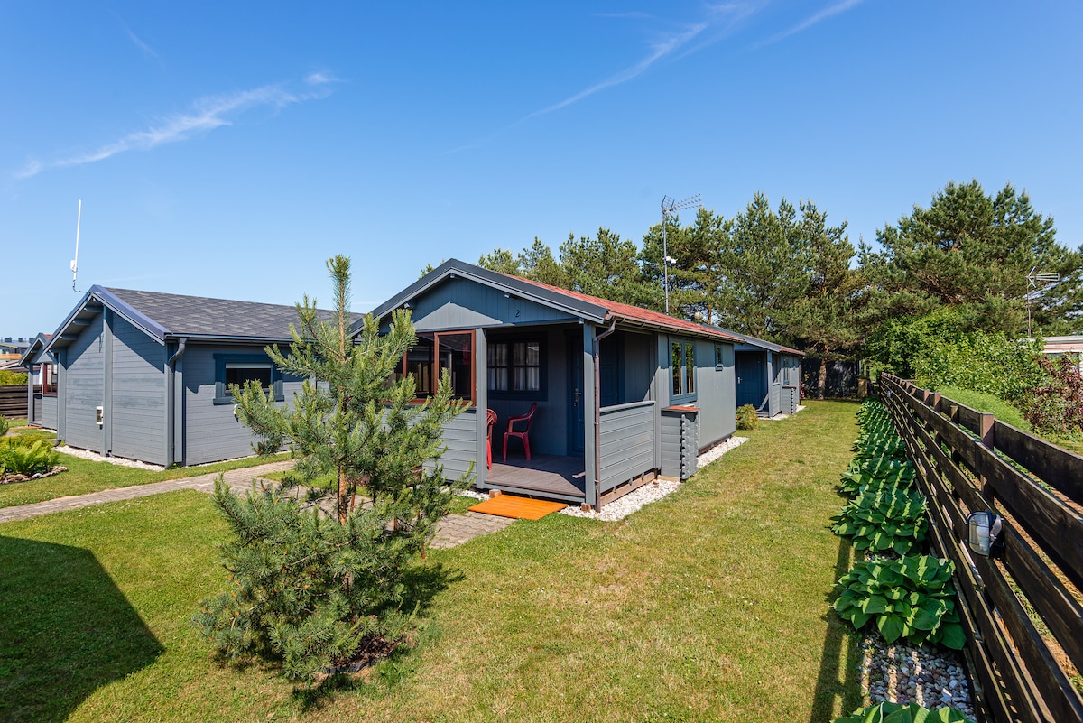 A row of wooden houses is set in a green grassy area, surrounded by trees and shrubs. Each house features a welcoming entrance with a small terrace, complemented by outdoor seating. The clear blue sky enhances the peaceful atmosphere of the surroundings.