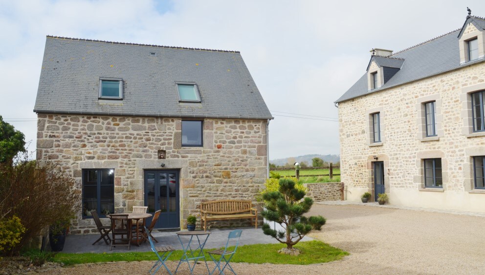 A charming stone house features a gravel courtyard, showcasing a wooden bench and a small dining set. Large windows allow natural light to brighten the interior. Surrounding greenery adds a touch of nature to the peaceful setting.