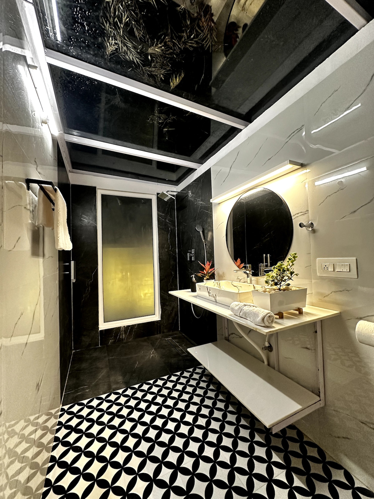 A stylish bathroom features dark marble walls and contrasting white fixtures. A circular mirror is mounted above a sleek countertop, which includes small potted plants. Natural light filters in through a skylight, illuminating the patterned black and white floor tiles. Towel racks are neatly positioned.