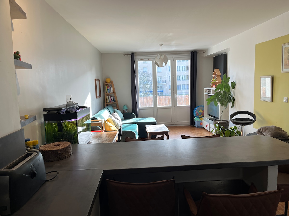 A spacious living area features large windows that flood the room with natural light, creating a bright and open atmosphere. A comfortable sofa and a small coffee table are positioned conveniently. A kitchenette is partially visible, with modern appliances and a bar counter with seating.