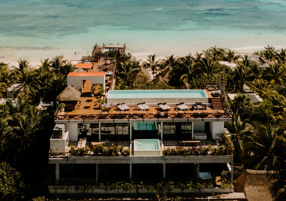 An aerial view captures the hotel's layout, showing multiple terraces and a central pool surrounded by sun loungers. Lush palm trees frame the property, and the turquoise waters of the Caribbean are visible in the background, enhancing the tranquil coastal setting.