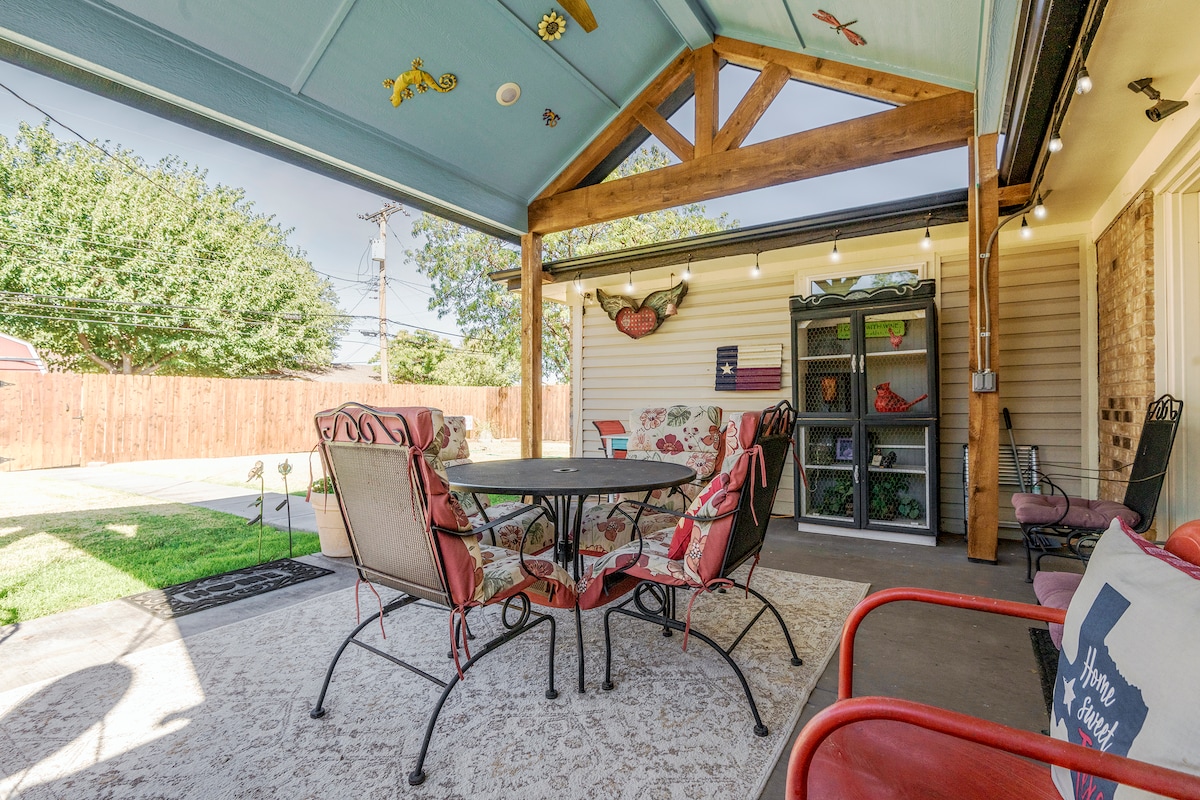 A covered patio space features a round table surrounded by four comfortable chairs with floral cushions. Soft lighting hangs overhead, creating a cozy ambiance. A patterned rug covers the patio floor, and a tidy yard with greenery is visible beyond a privacy fence.