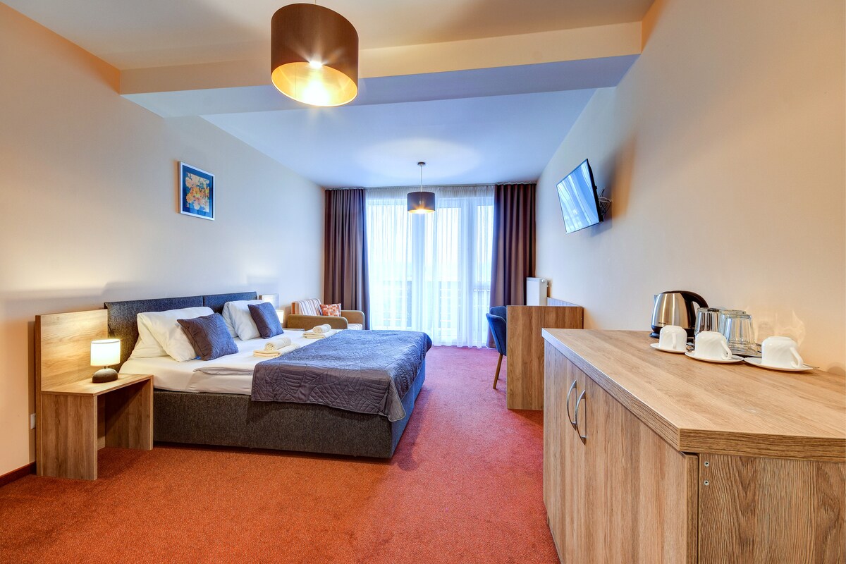 A spacious room contains two beds with soft bedding. A comfortable seating area is provided near a large window, allowing natural light to brighten the interior. A flat-screen TV is mounted on the wall, and a wooden cabinet displays a kettle and cups.