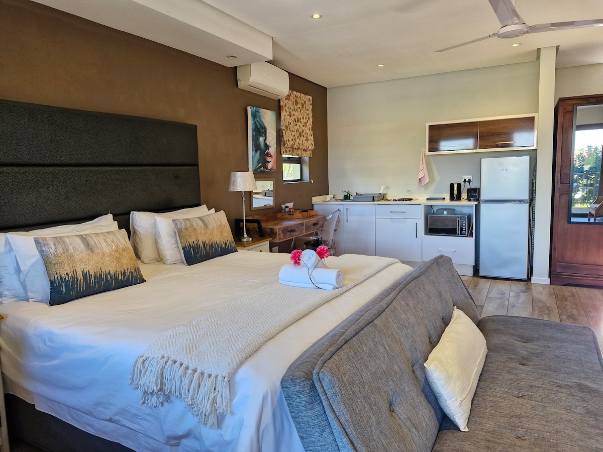 A spacious room features a comfortable king size bed with decorative pillows, accompanied by a double sleeper couch. A kitchenette with modern appliances is visible, along with a dining area. Natural light enters through windows, and air conditioning is available.