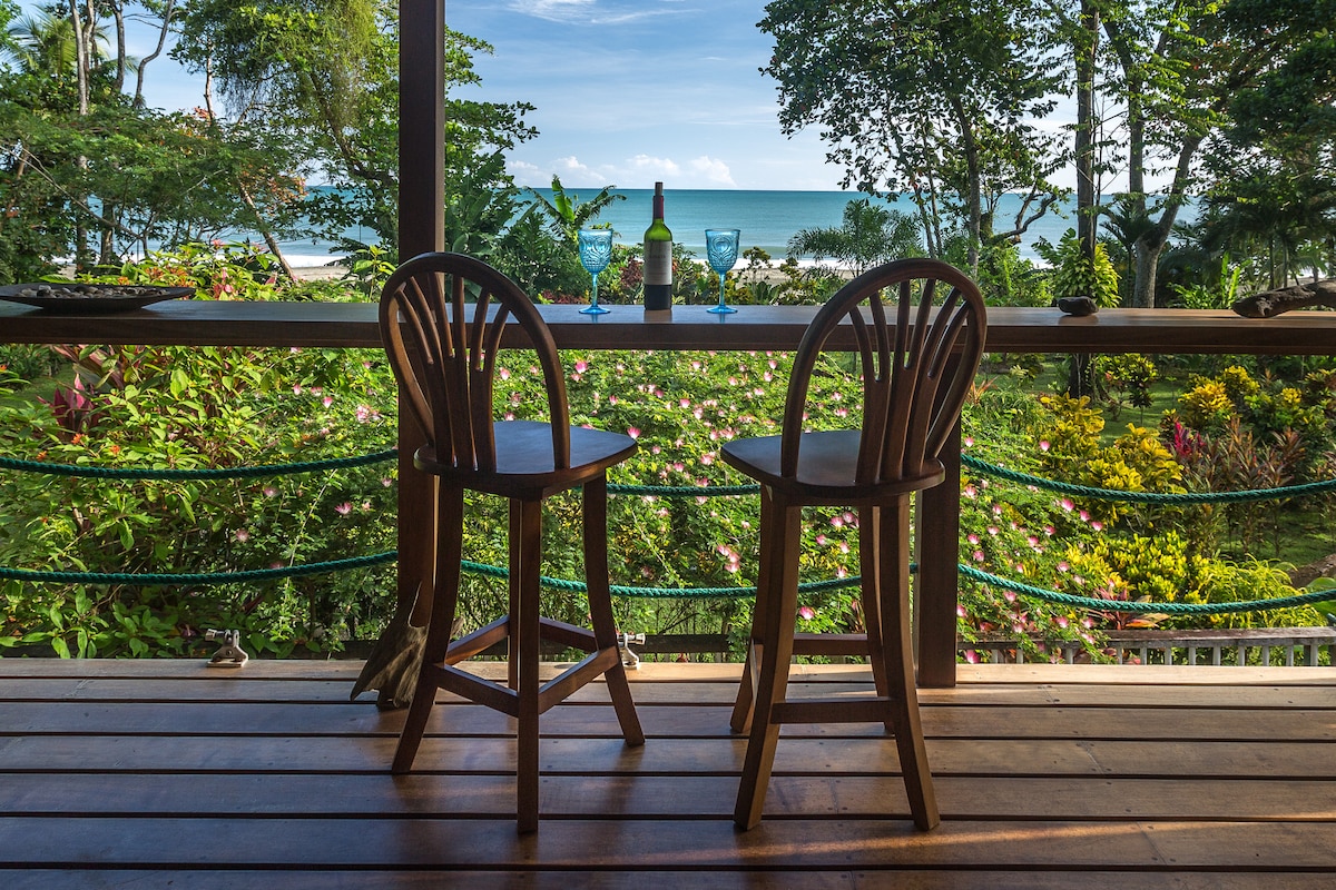 Two wooden bar stools are positioned on a wooden veranda, overlooking a vibrant garden that leads to the ocean. A bottle and two glasses sit on the table, with the blue sea and sky visible in the background, framed by lush greenery.