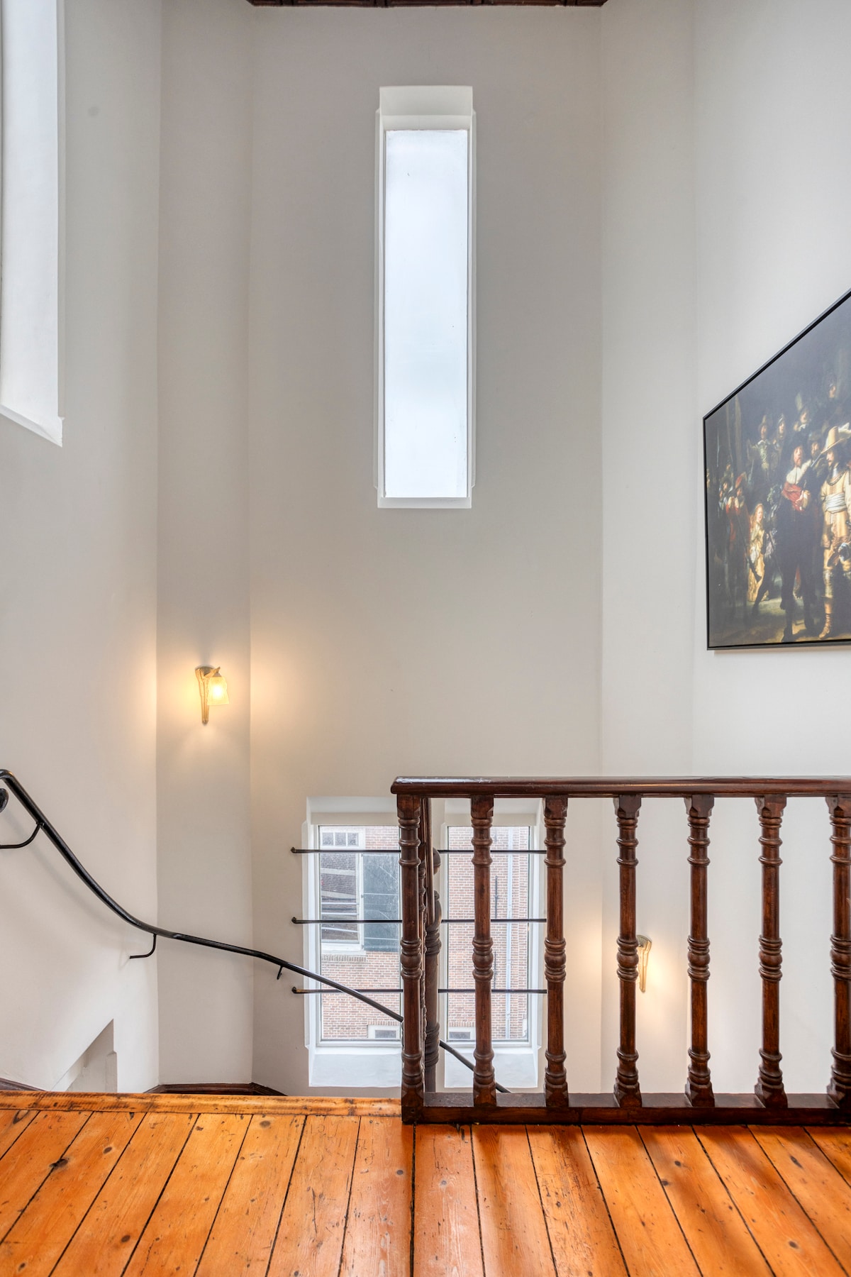 Stairs are featured within a well-lit space, complemented by a tall window allowing natural light to filter in. A wooden railing provides a classic touch, while the warm wood flooring adds warmth to the area. A large painting is displayed along one wall.