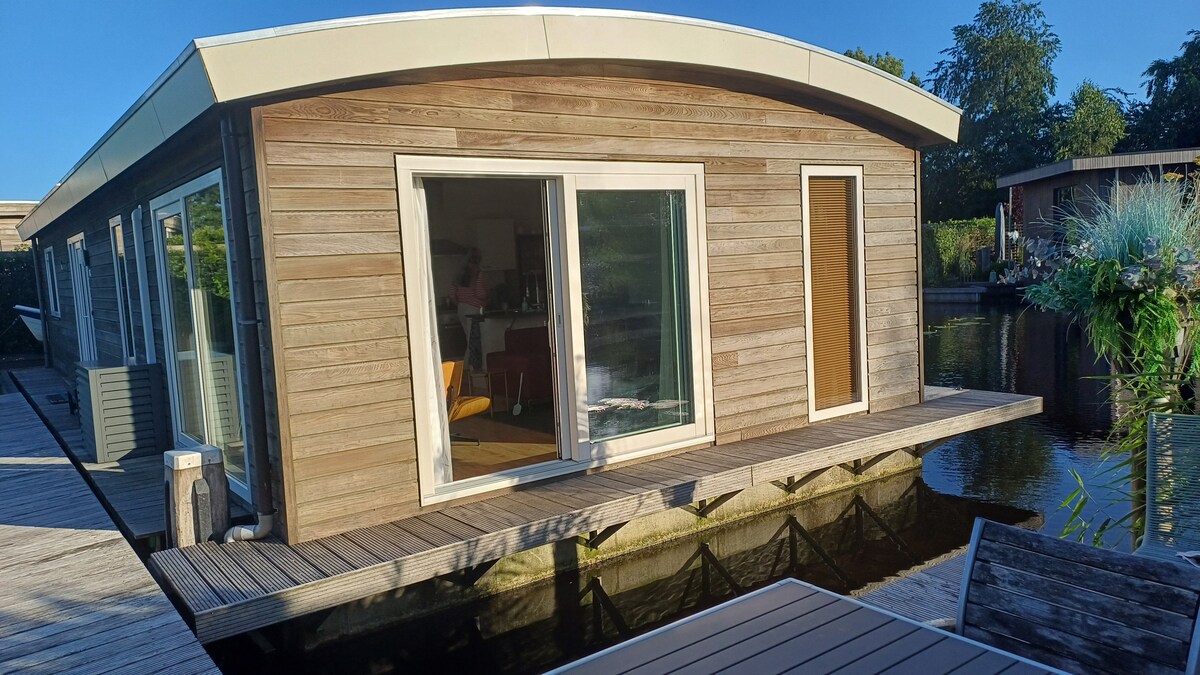 A modern houseboat is depicted, showcasing a sleek wooden exterior with large glass windows. The structure extends over the water, with a walkway leading to it. The setting is surrounded by greenery, creating a serene and private atmosphere.