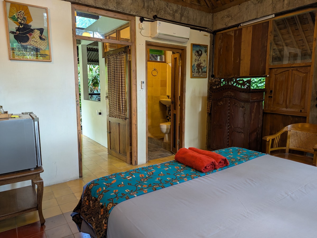 A spacious bedroom features a comfortable bed draped in a vibrant blanket, with a red towel neatly placed on top. Natural light filters through the open doorways, highlighting the rustic wood furniture and cool tile flooring. An ensuite bathroom is visible in the background.