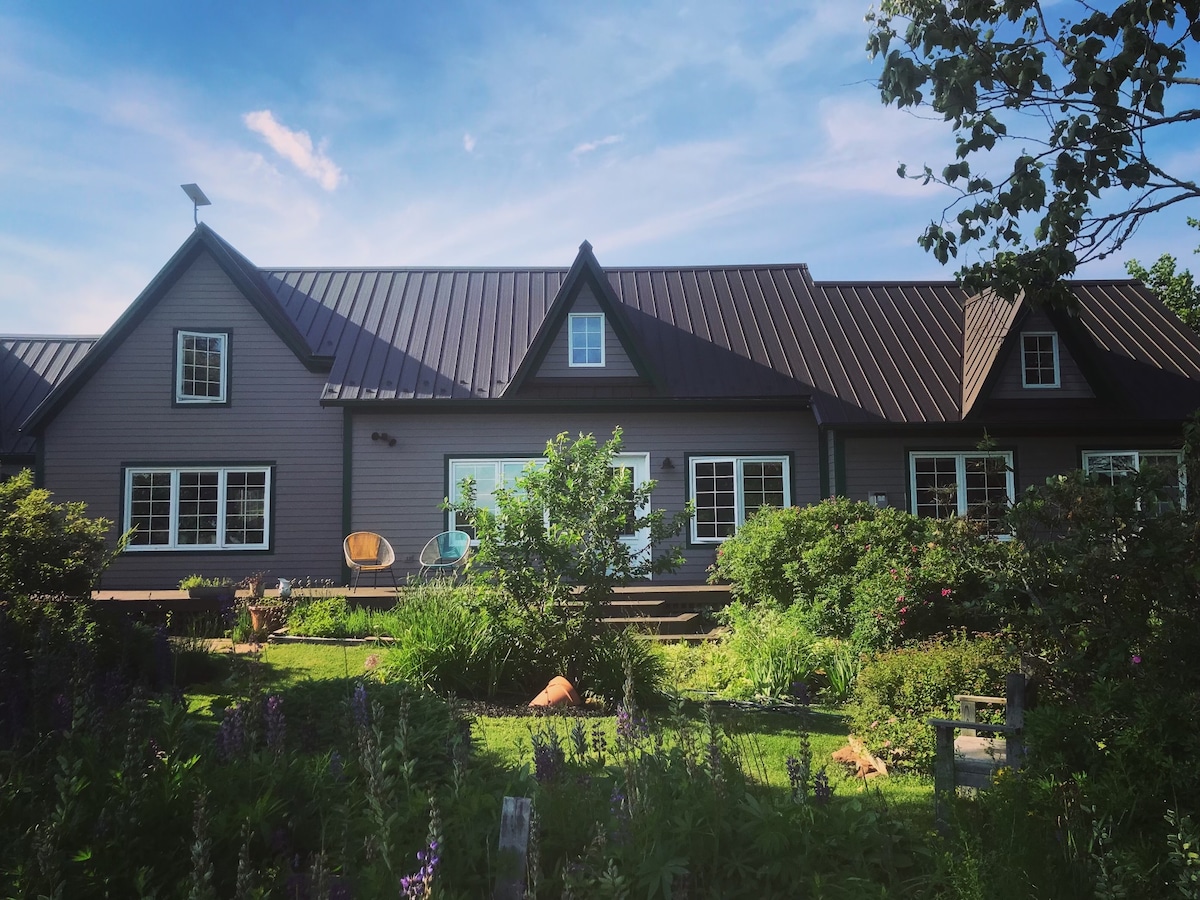 The exterior of The Cottage is set amidst lush greenery and vibrant flowers, featuring a well-maintained garden. The house boasts a sloped roof with multiple gables, large windows, and a welcoming deck area, inviting relaxation in a serene natural setting.