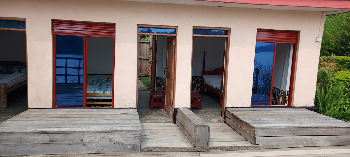 A welcoming accommodation structure with multiple entries. Each room features large windows that provide views of the surrounding landscape and lake. Wooden decks lead to the entrances, creating accessible access to each room. The interior spaces are simple, reflecting comfort and functionality.