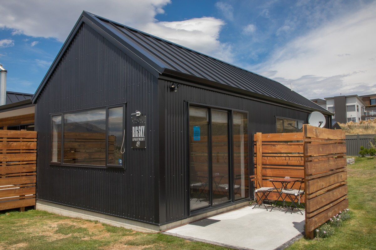 The exterior of the apartment is presented, showcasing a sleek black building with a metal roof. Large windows allow natural light inside. A small outdoor area features a table and chairs, surrounded by wooden fencing, creating a sense of privacy.