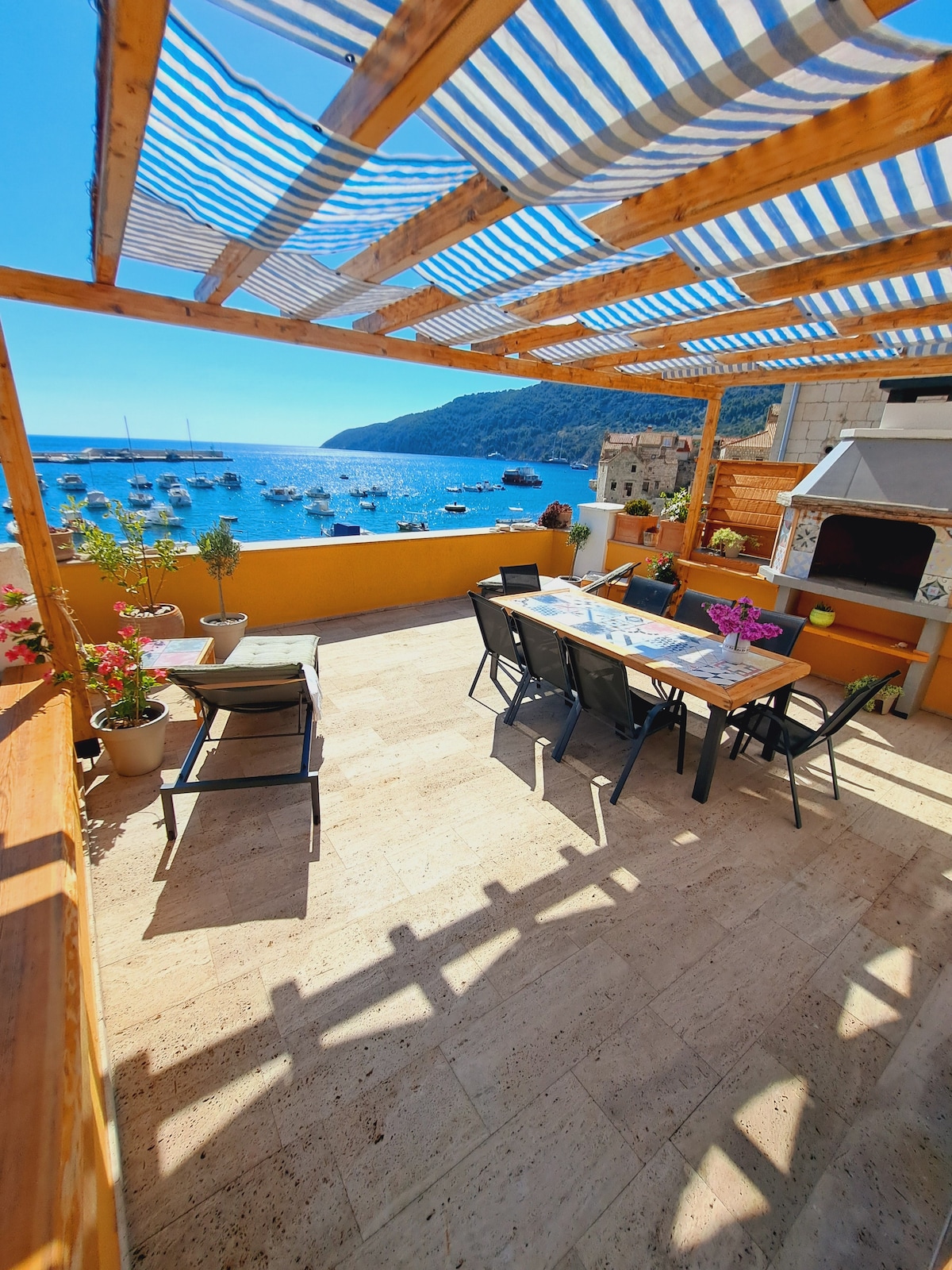 An expansive terrace features a dining area with a long table and chairs, positioned under a striped awning. A grill is located nearby, with a backdrop of clear blue waters and boats in the harbor. Potted flowers add a touch of greenery to the inviting outdoor space.