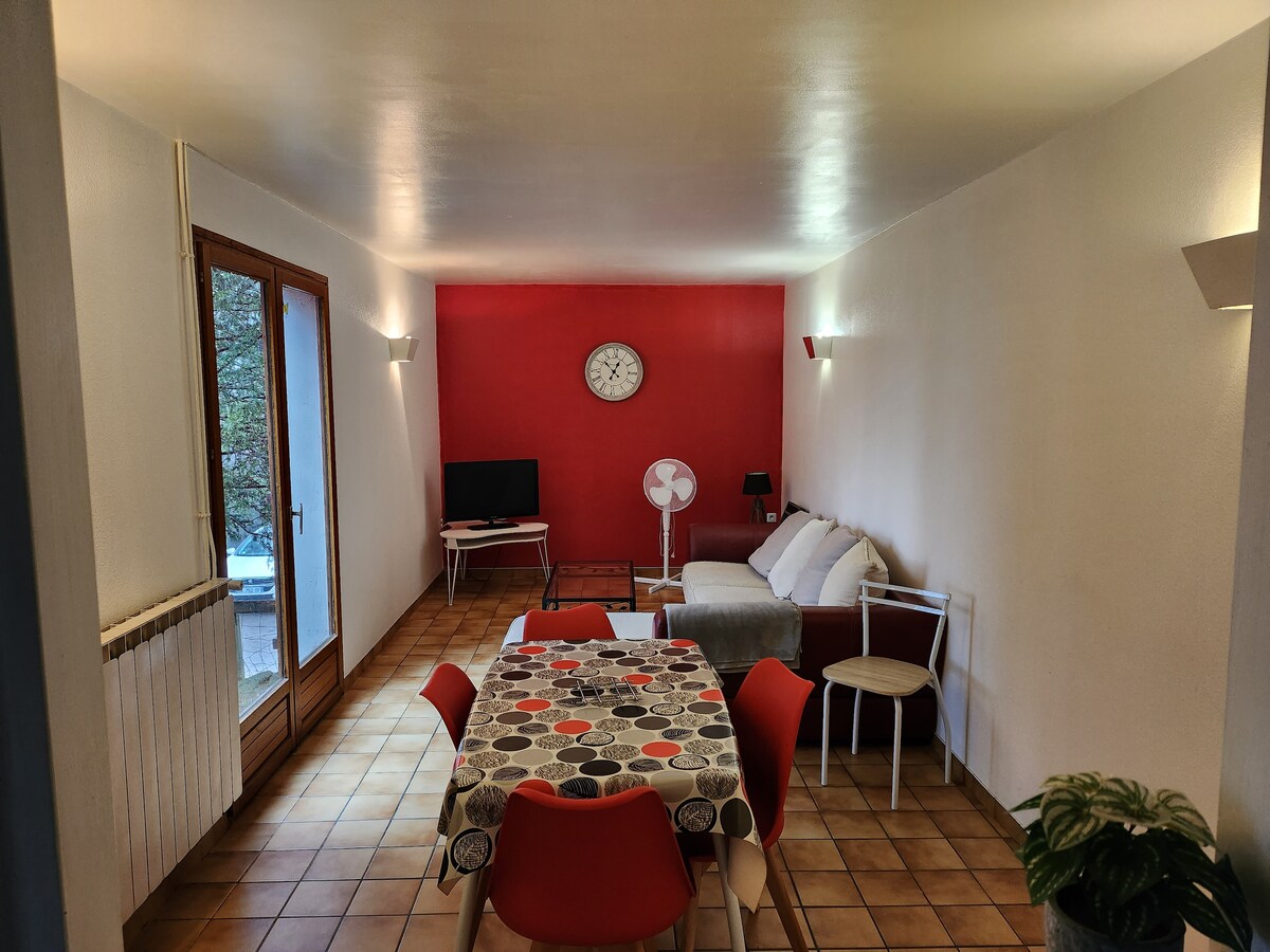A spacious living area features a red accent wall and tile flooring. A round dining table is surrounded by red chairs. A comfortable sofa sits across from a flat-screen TV, with a clock mounted on the wall and a fan nearby, allowing for a relaxing atmosphere.