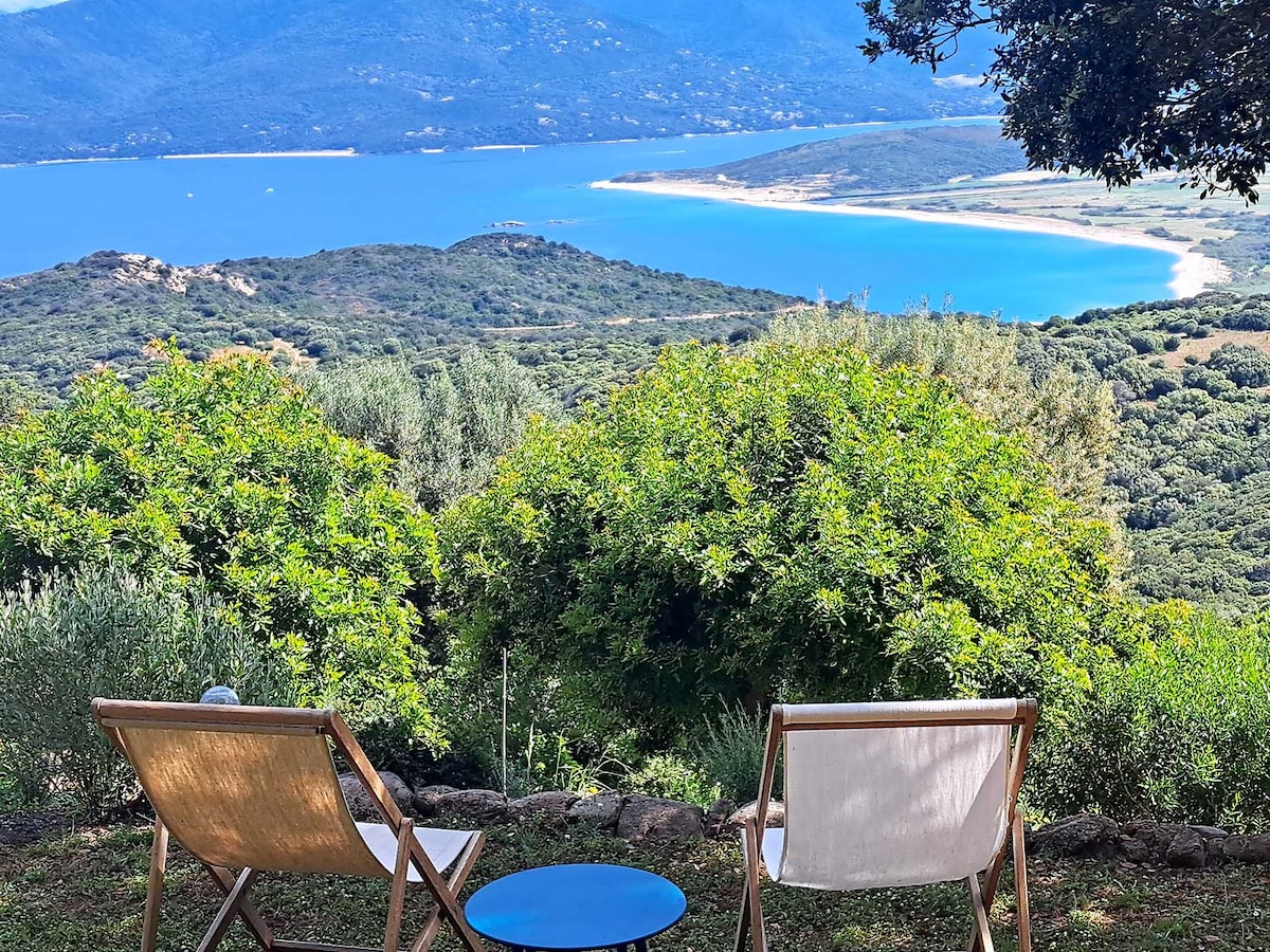 Two wooden chairs face a stunning coastal view, framed by lush greenery. A small blue table is positioned between them, creating a serene spot to enjoy the landscape of white sand beaches and distant mountains under a clear sky.
