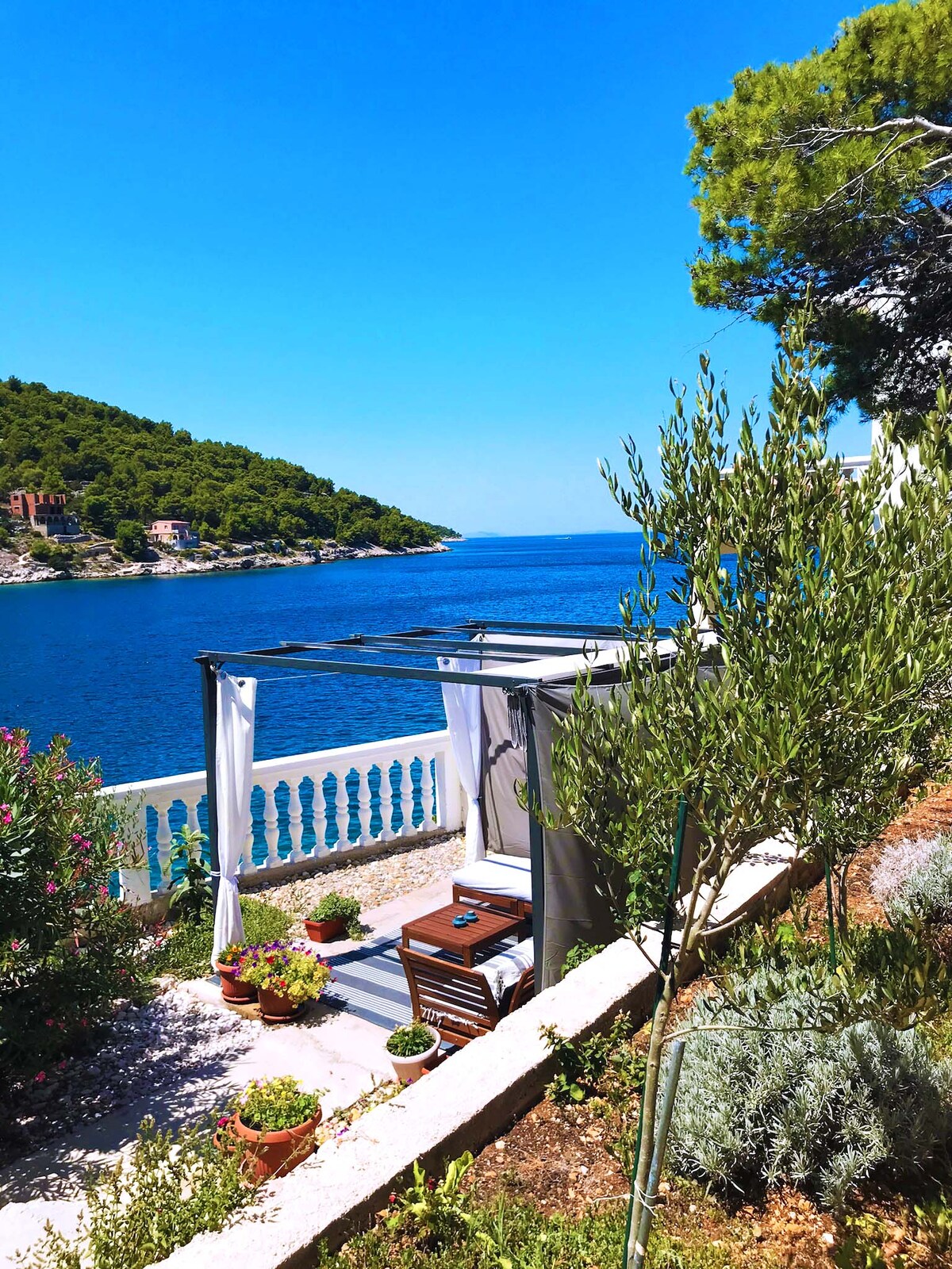 A serene outdoor space features a shaded seating area with a small table and two chairs, surrounded by lush greenery and potted plants. The clear blue sea can be seen in the background, framed by a hillside of trees, inviting relaxation by the water.