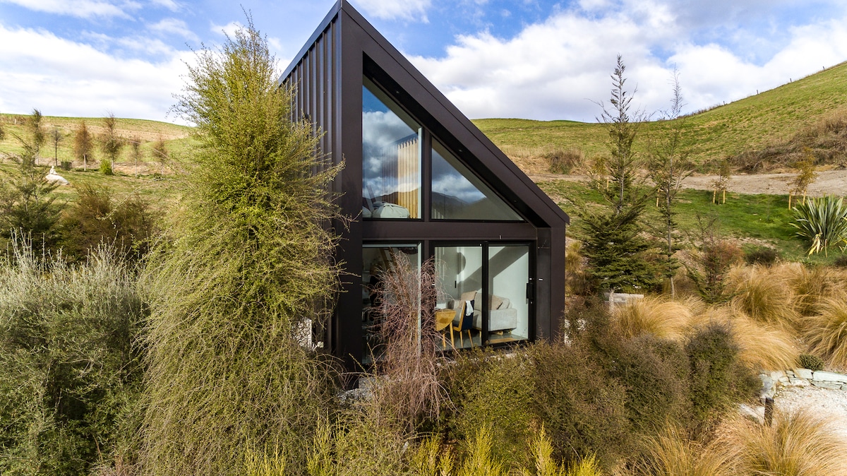 An architecturally designed tiny home is surrounded by lush greenery and native plants. The sloped roof adds character, while large floor-to-ceiling windows provide natural light and views of the landscape. The exterior blends modern design with natural elements, creating a peaceful retreat.