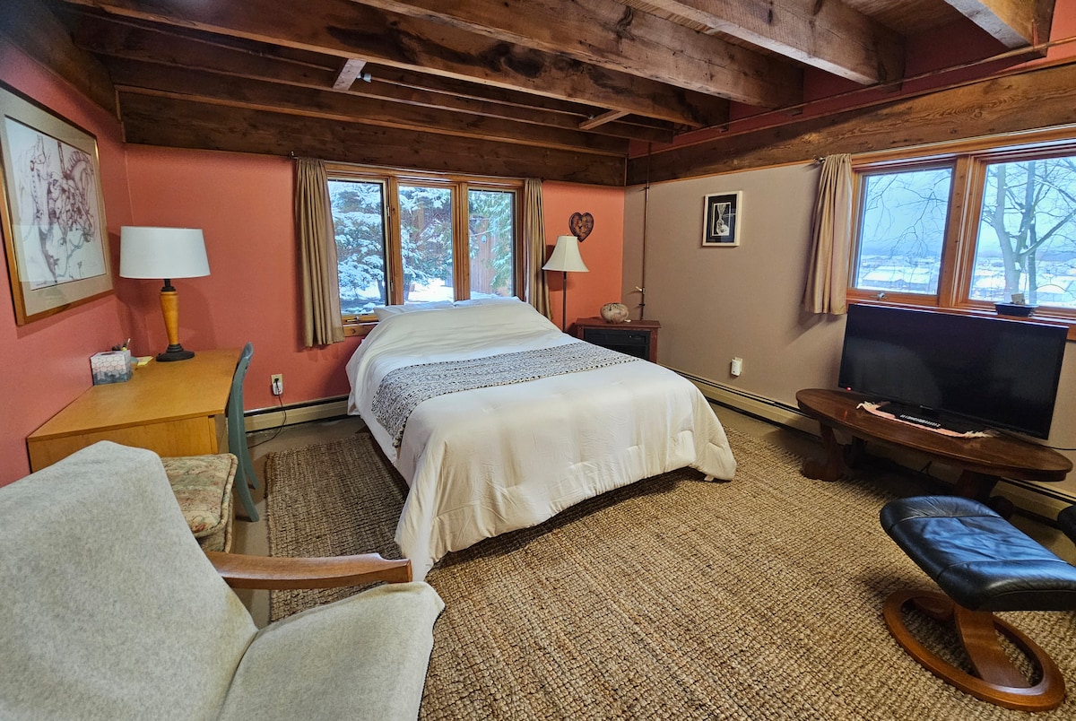 A spacious sleeping area features a large bed dressed in neutral bedding, complemented by wooden beams that enhance the rustic charm. A cozy armchair and a round table are positioned nearby. Large windows provide natural light and views of the outdoor space, with winter scenery visible.
