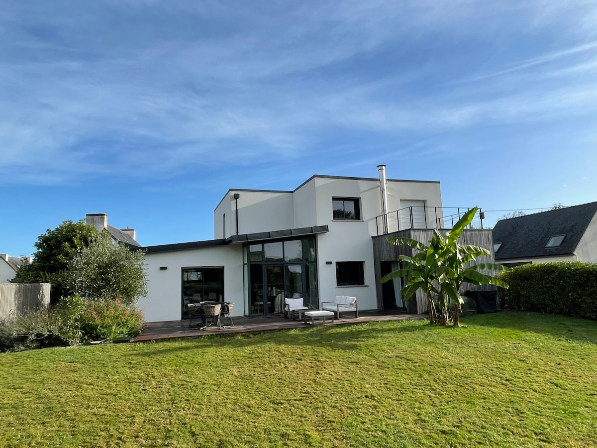 A modern two-story house is depicted, featuring large glass doors that open to a spacious wooden terrace. The lush green garden surrounds the property, with well-maintained plants and seating areas visible. A balcony on the upper level adds to the architectural appeal.