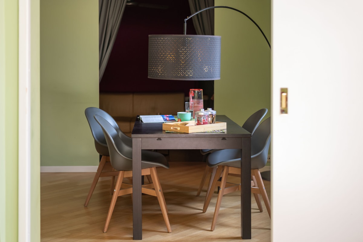 A dining area is visible with a modern table complemented by four chairs. A large, stylish lamp hangs overhead, providing focused lighting. Small decorative items adorn the table, and green walls create a subtle backdrop. The space leads to a cozy lounge area beyond.