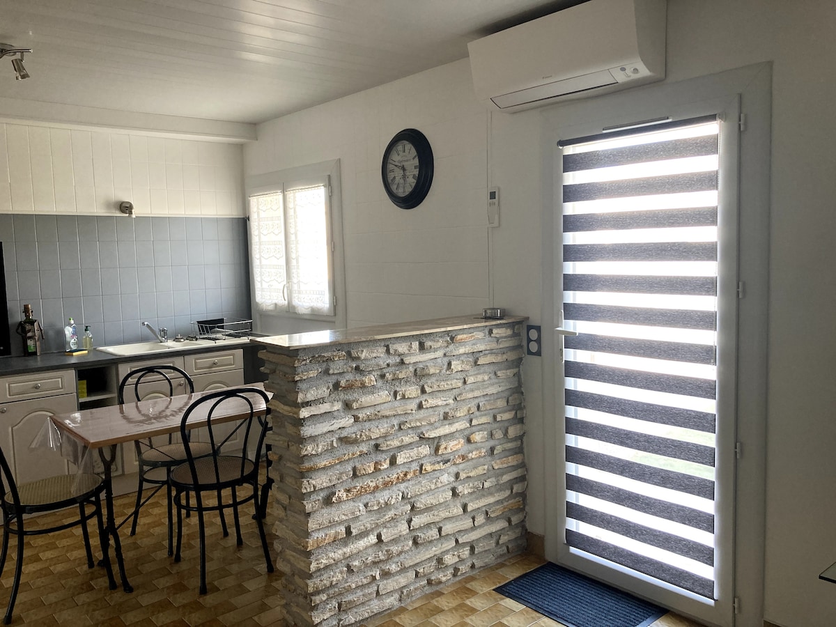 An open kitchen area features a stone bar with a dining table and black metal chairs. A wall clock is mounted above the bar, and natural light enters through a large door with horizontal stripes. A shaded indoor environment is created by the air conditioning unit.