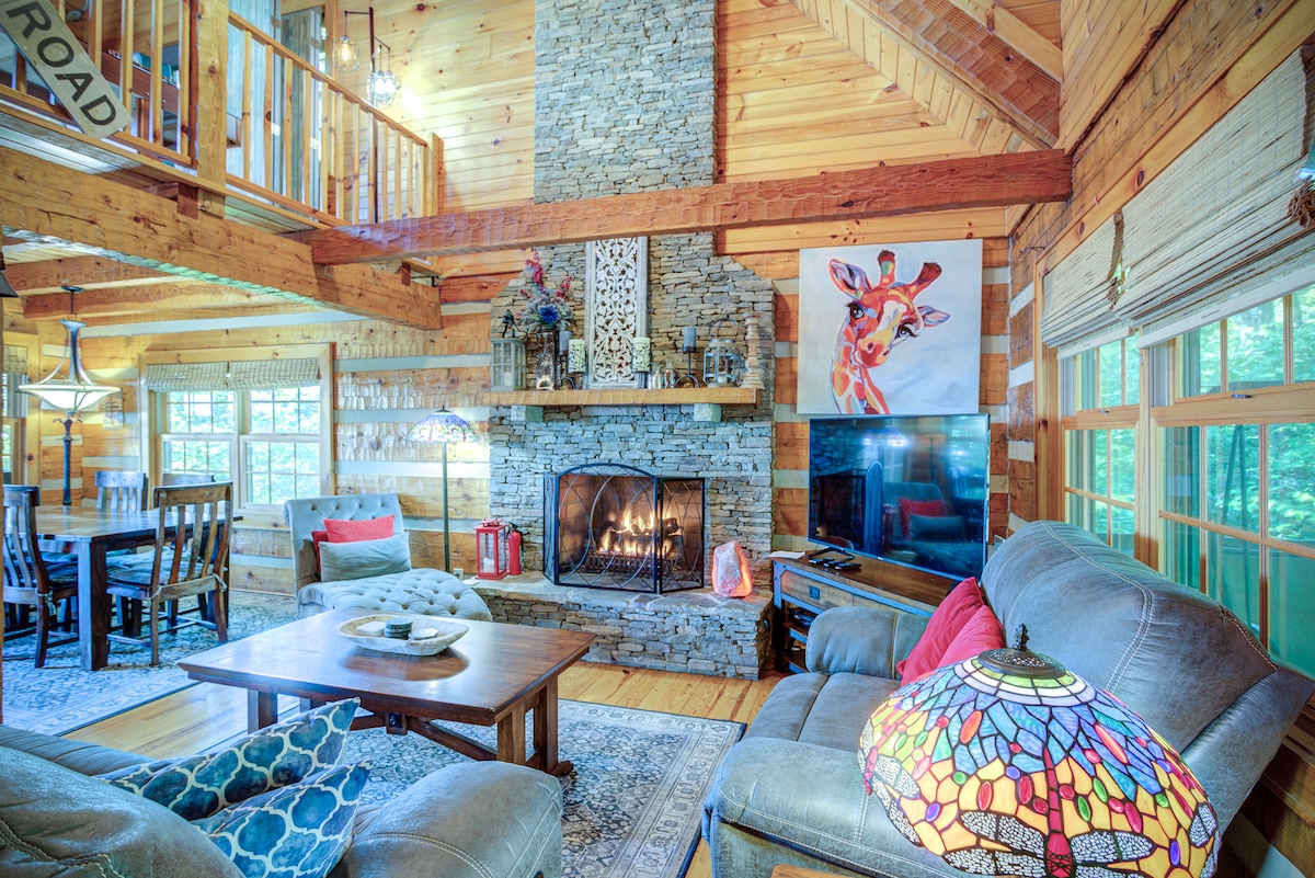 A cozy living area features a stone fireplace as its focal point, surrounded by wooden walls and tasteful decor. Comfortable seating is provided by a large sectional sofa and a lounge chair. A colorful stained glass lamp adds a decorative touch, with a view of the dining area nearby.