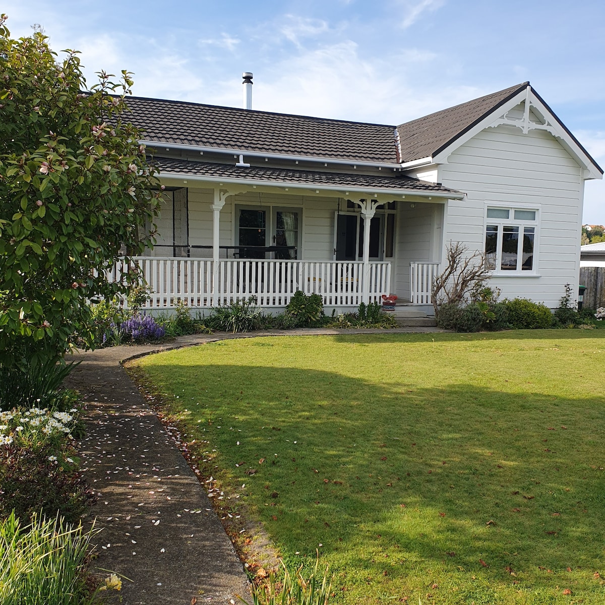 A charming white house is set against a clear blue sky, featuring a welcoming porch and expansive front lawn. Neatly trimmed grass and scattered flower beds enhance the outdoor space, leading to a paved pathway that invites exploration.