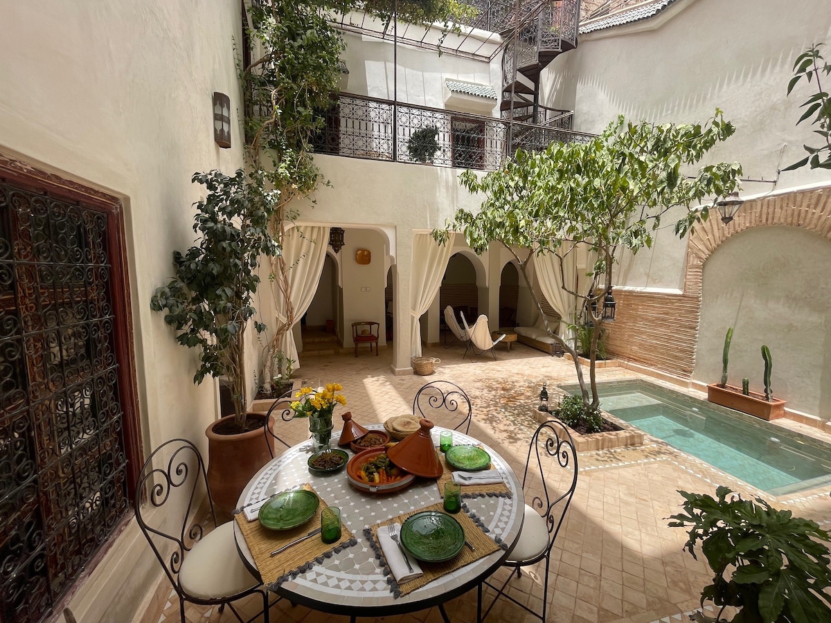A tranquil patio is highlighted by a small pool and lush greenery, including a tree and potted plants. A round table set for dining features colorful plates and a vase of flowers, complemented by soft drapes that create an inviting atmosphere.