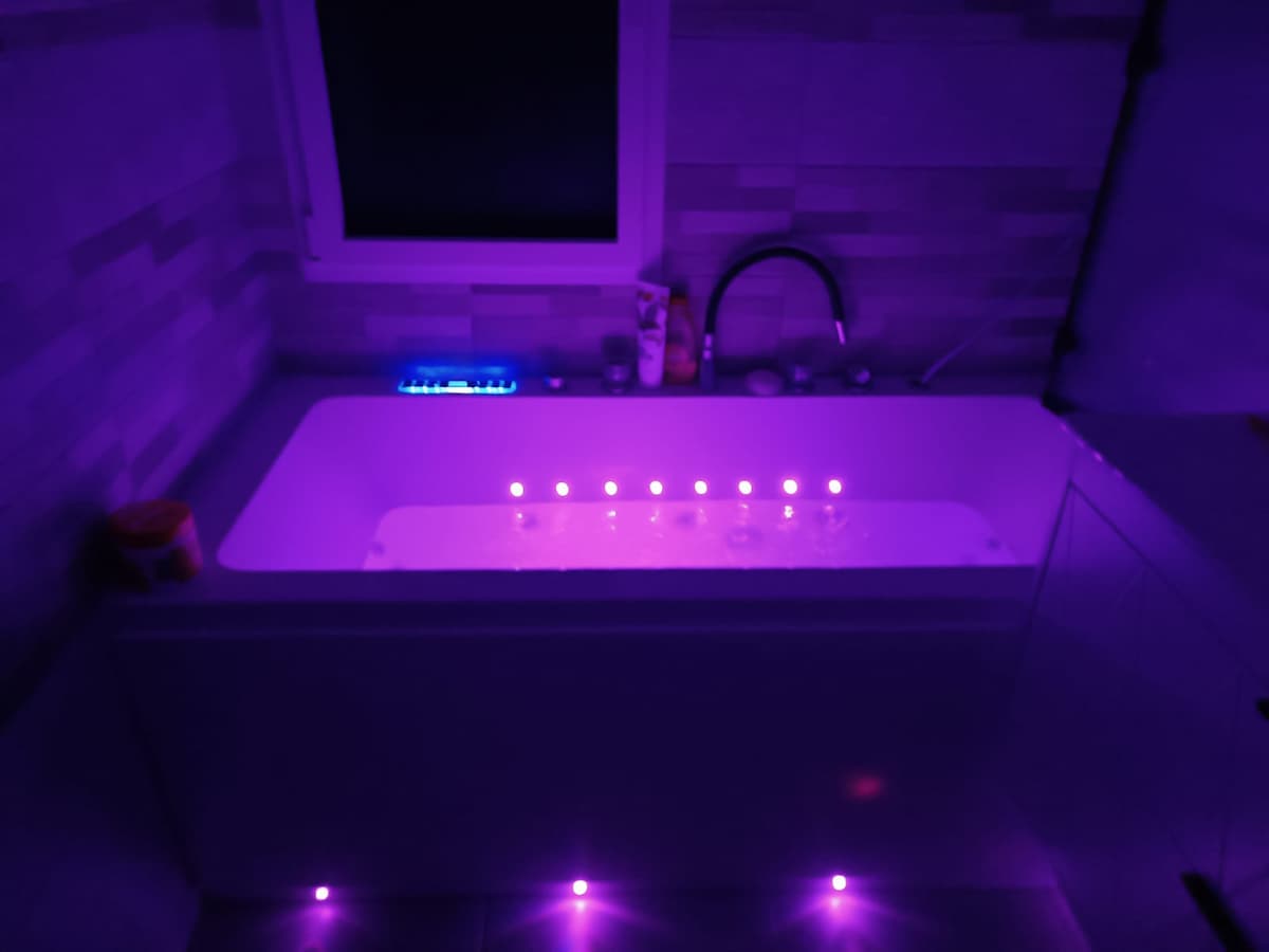 A modern bathroom features a sleek bathtub illuminated with soft purple lighting, creating a calming ambiance. The surrounding walls showcase a stylish tiled design, and a window allows natural light to enter. Various toiletries are visible on the edge of the bathtub.