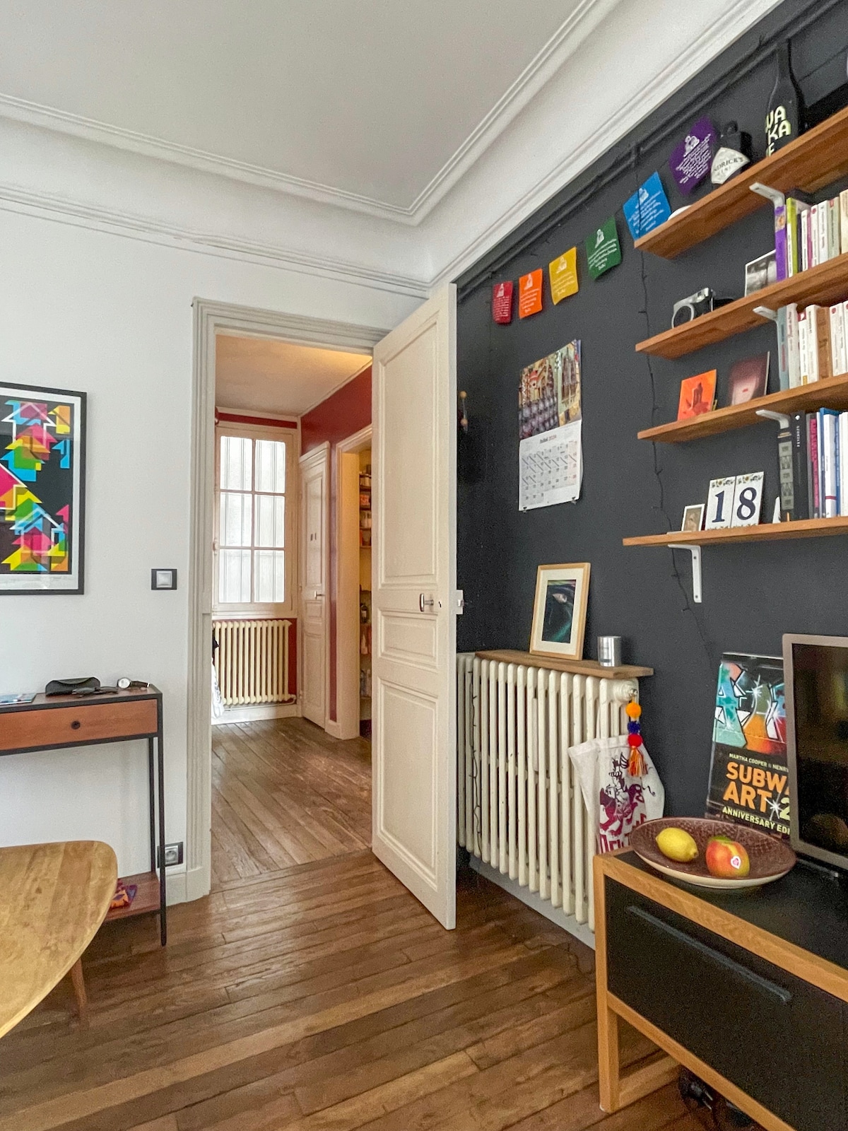 A modern entrance area features a tasteful combination of dark and light colors. A wooden table sits beside a wall adorned with shelves and colorful artwork, while natural light fills the space through a nearby window. The flooring consists of classic wooden planks.